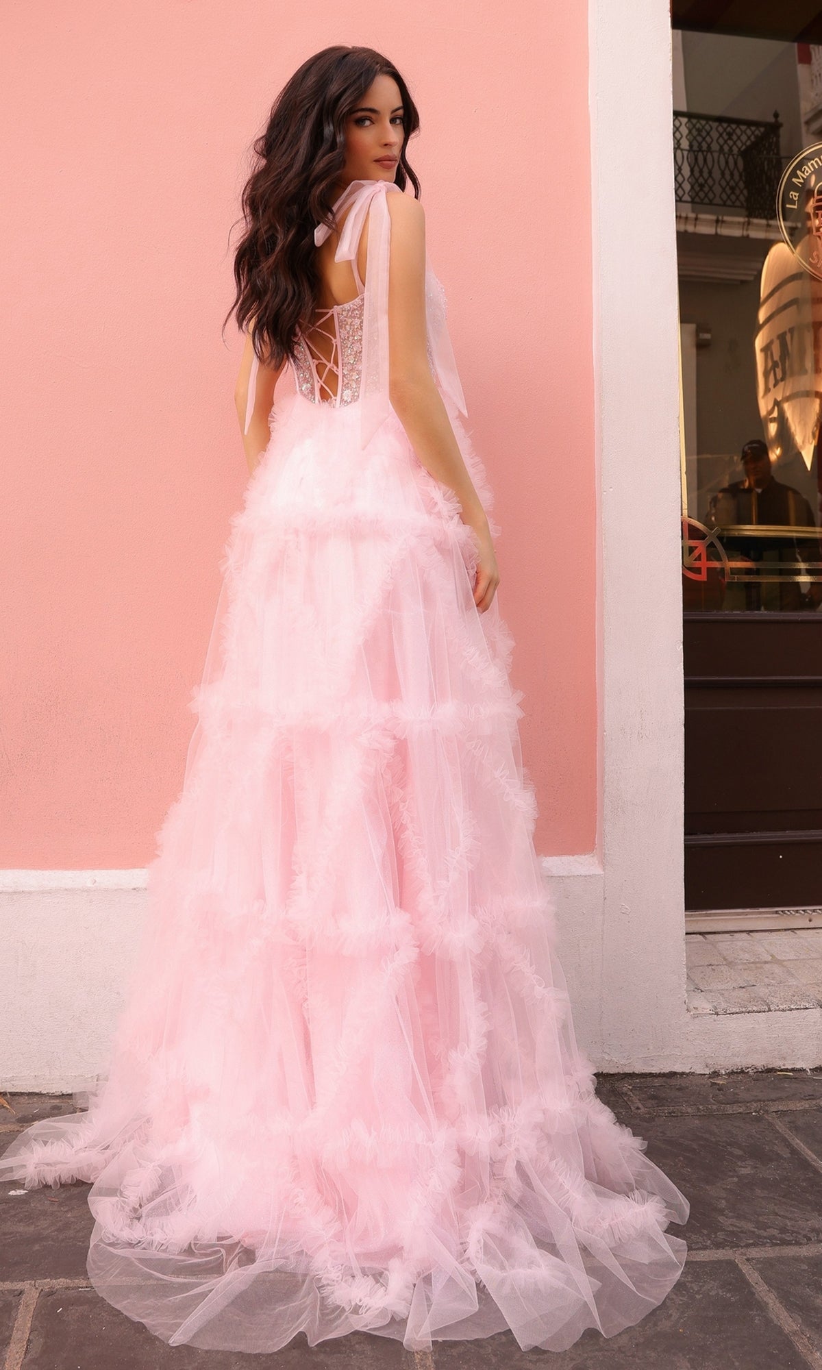 A woman in the Shoulder-Bows Long Ruffled Prom Gown T1340, a long light pink tulle dress with a beaded corset bodice and lace-up back, stands outdoors against a pale pink wall, glancing over her shoulder.